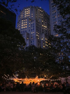 An evening scene features a brightly lit outdoor gathering beneath trees adorned with string lights. In the background, a tall building with numerous illuminated windows towers under a deep blue sky. People are seated and mingling under a tent, contributing to a lively yet relaxed atmosphere.