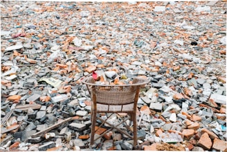 A wicker chair is situated in the foreground, placed on a vast expanse of rubble and debris. The ground is covered with broken bricks, stones, and scattered trash, creating a chaotic and desolate scene.