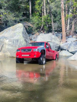 Shiny red pickup truck loaded with camping gear parked by a forest lake.