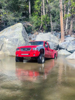 Shiny red pickup truck loaded with camping gear parked by a forest lake.