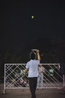 A person is playing badminton outside at night. The shuttlecock is in mid-air, and the player is poised to hit it with a racket. A sports net is set up between two poles, and another person stands on the opposite side witnessing or waiting. A motorcycle is parked nearby, and the background is dark, suggesting evening or night time.
