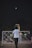 A person is playing badminton outside at night. The shuttlecock is in mid-air, and the player is poised to hit it with a racket. A sports net is set up between two poles, and another person stands on the opposite side witnessing or waiting. A motorcycle is parked nearby, and the background is dark, suggesting evening or night time.