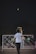 A person is playing badminton outside at night. The shuttlecock is in mid-air, and the player is poised to hit it with a racket. A sports net is set up between two poles, and another person stands on the opposite side witnessing or waiting. A motorcycle is parked nearby, and the background is dark, suggesting evening or night time.