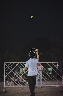 A person is playing badminton outside at night. The shuttlecock is in mid-air, and the player is poised to hit it with a racket. A sports net is set up between two poles, and another person stands on the opposite side witnessing or waiting. A motorcycle is parked nearby, and the background is dark, suggesting evening or night time.