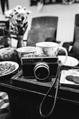 A black-and-white image depicting a vintage camera placed on a wooden table surrounded by assorted ceramic cups and plates. Behind the camera are blurred chairs and a vase with flowers.