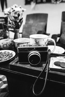 A black-and-white image depicting a vintage camera placed on a wooden table surrounded by assorted ceramic cups and plates. Behind the camera are blurred chairs and a vase with flowers.