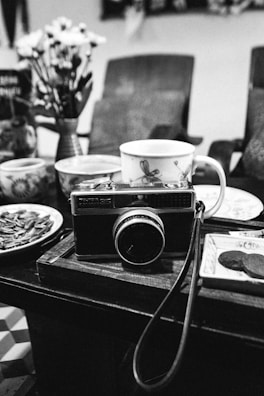 A black-and-white image depicting a vintage camera placed on a wooden table surrounded by assorted ceramic cups and plates. Behind the camera are blurred chairs and a vase with flowers.