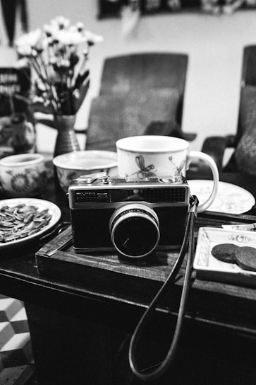 A black-and-white image depicting a vintage camera placed on a wooden table surrounded by assorted ceramic cups and plates. Behind the camera are blurred chairs and a vase with flowers.