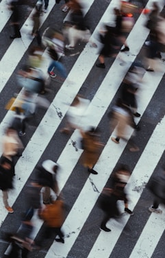 Blurred motion captures a busy street crossing with numerous pedestrians in mid-step, walking in various directions across marked crosswalk lines.