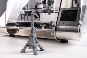 A 3D printer is producing a miniature gray model of the Eiffel Tower. The printer has a sleek design with a metallic surface and a transparent casing that reveals the internal mechanics.