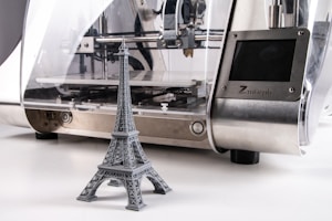 A 3D printer is producing a miniature gray model of the Eiffel Tower. The printer has a sleek design with a metallic surface and a transparent casing that reveals the internal mechanics.