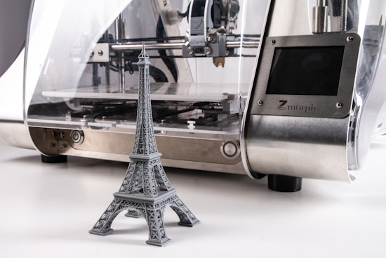 A 3D printer is producing a miniature gray model of the Eiffel Tower. The printer has a sleek design with a metallic surface and a transparent casing that reveals the internal mechanics.