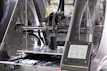 A 3D printer in action, with a partially completed black object on the print bed. The machine features transparent casing, metal components, and a control panel displaying settings and progress information.