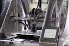 A 3D printer in action, with a partially completed black object on the print bed. The machine features transparent casing, metal components, and a control panel displaying settings and progress information.