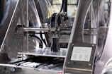 A 3D printer in action, with a partially completed black object on the print bed. The machine features transparent casing, metal components, and a control panel displaying settings and progress information.