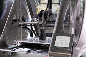 A 3D printer in action, with a partially completed black object on the print bed. The machine features transparent casing, metal components, and a control panel displaying settings and progress information.