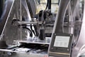 A 3D printer in action, with a partially completed black object on the print bed. The machine features transparent casing, metal components, and a control panel displaying settings and progress information.