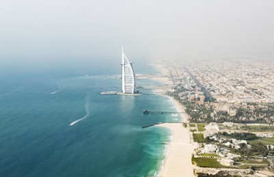 Aerial view of a modern, sail-shaped building situated on an artificial island, surrounded by the turquoise waters of the sea. A wide expanse of cityscape extends along the coastline, with a sandy beach leading towards the urban area.