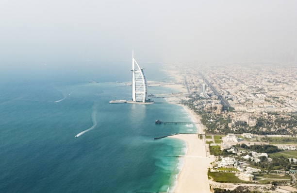 Aerial view of a modern, sail-shaped building situated on an artificial island, surrounded by the turquoise waters of the sea. A wide expanse of cityscape extends along the coastline, with a sandy beach leading towards the urban area.