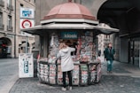 A street kiosk is stocked with various colorful magazines and newspapers, attended by a person in a gray coat who is browsing the selection. The kiosk is located on a cobblestone street, surrounded by historic buildings. In the background, a pedestrian is walking towards the camera.