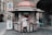 A street kiosk is stocked with various colorful magazines and newspapers, attended by a person in a gray coat who is browsing the selection. The kiosk is located on a cobblestone street, surrounded by historic buildings. In the background, a pedestrian is walking towards the camera.