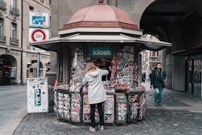 A street kiosk is stocked with various colorful magazines and newspapers, attended by a person in a gray coat who is browsing the selection. The kiosk is located on a cobblestone street, surrounded by historic buildings. In the background, a pedestrian is walking towards the camera.