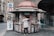 A street kiosk is stocked with various colorful magazines and newspapers, attended by a person in a gray coat who is browsing the selection. The kiosk is located on a cobblestone street, surrounded by historic buildings. In the background, a pedestrian is walking towards the camera.