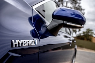 A close-up side view of a blue hybrid car, focusing on the side mirror and the 'HYBRID' badge. The background is slightly blurred, highlighting the details of the vehicle.