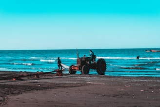 On a beach with dark sand, a tractor is positioned near the shoreline. A person stands near the tractor, and another is seated at the controls. The ocean waves roll gently in the background, and the sky is a clear shade of blue. Fishing boats can be seen in the distance.