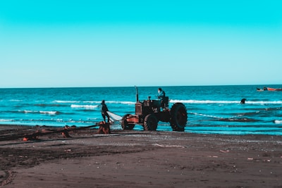 On a beach with dark sand, a tractor is positioned near the shoreline. A person stands near the tractor, and another is seated at the controls. The ocean waves roll gently in the background, and the sky is a clear shade of blue. Fishing boats can be seen in the distance.