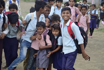 A group of school children are gathered closely, some smiling and others laughing. They are wearing school uniforms with backpacks, indicating they are either going to or coming from school. The background shows a dirt ground and more students in the distance, suggesting an outdoor setting, possibly a school yard.