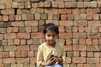 A young girl with dark hair sits in front of a wall made of stacked bricks. She is wearing a light-colored shirt and holding a black object in her hands, while looking directly at the camera.