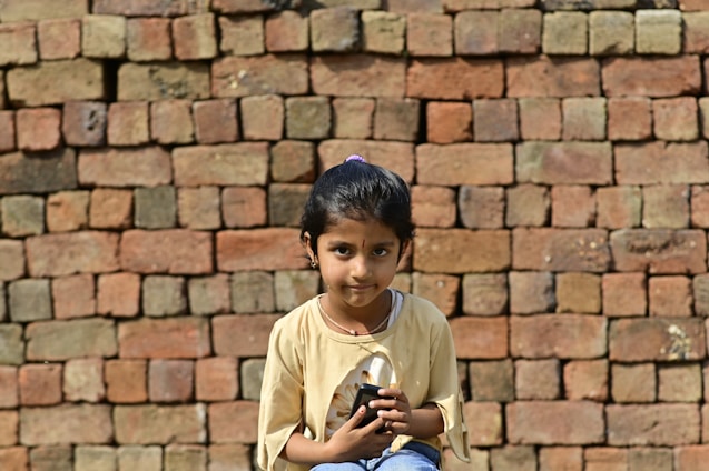 A young girl with dark hair sits in front of a wall made of stacked bricks. She is wearing a light-colored shirt and holding a black object in her hands, while looking directly at the camera.