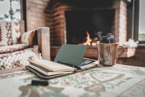 A cozy indoor scene of a wooden table with an open notebook and a pen resting on it. Beside them, a steaming striped mug sits on the table. In the background, a fireplace with a crackling fire is set into a brick wall. A crocheted blanket is draped over the arm of a wooden chair, contributing to the warm and inviting atmosphere.