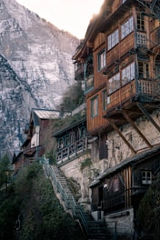 Rustic wooden houses are nestled on a steep hillside, with intricate balconies overlooking a dramatic mountainous backdrop. Snow-capped peaks surround the scene, and a stone staircase weaves through lush greenery, adding a quaint charm.