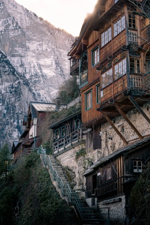Rustic wooden houses are nestled on a steep hillside, with intricate balconies overlooking a dramatic mountainous backdrop. Snow-capped peaks surround the scene, and a stone staircase weaves through lush greenery, adding a quaint charm.