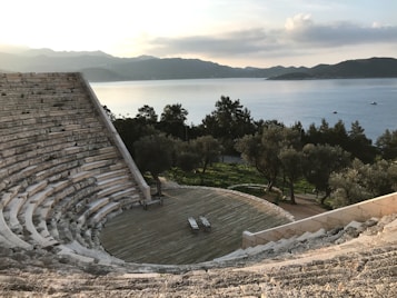 An ancient stone amphitheater with semicircular seating overlooks a scenic view of calm waters and distant mountains under a partly cloudy sky. Olive trees surround the space, adding greenery to the tranquil setting.
