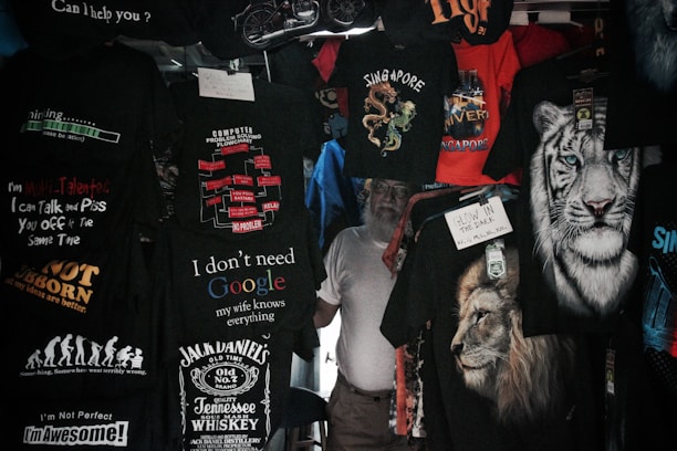 A variety of T-shirts are displayed with humorous and bold text designs, featuring phrases like 'I don't need Google,' and graphics such as lions and tigers. The T-shirts are predominantly dark with bright text and images.
