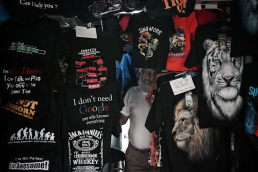 A variety of T-shirts are displayed with humorous and bold text designs, featuring phrases like 'I don't need Google,' and graphics such as lions and tigers. The T-shirts are predominantly dark with bright text and images.