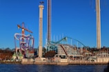 An amusement park with multiple rides including a roller coaster, a tall drop tower, and a colorful, winding track structure is situated by the water. The structures stand against a clear blue sky, and some trees and buildings can be seen in the background.