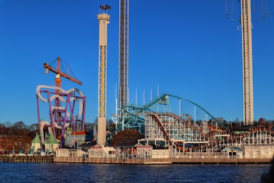 An amusement park with multiple rides including a roller coaster, a tall drop tower, and a colorful, winding track structure is situated by the water. The structures stand against a clear blue sky, and some trees and buildings can be seen in the background.