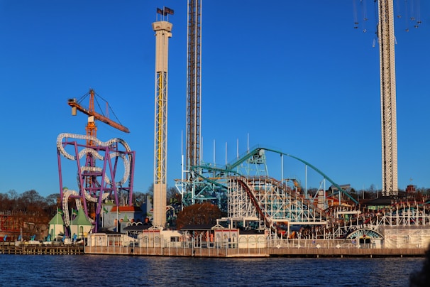 An amusement park with multiple rides including a roller coaster, a tall drop tower, and a colorful, winding track structure is situated by the water. The structures stand against a clear blue sky, and some trees and buildings can be seen in the background.