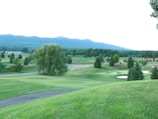 A lush, expansive golf course set against a backdrop of rolling hills and dense trees. The landscape features well-maintained grass, sand bunkers, and scattered trees, creating a serene and picturesque setting.