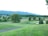 A lush, expansive golf course set against a backdrop of rolling hills and dense trees. The landscape features well-maintained grass, sand bunkers, and scattered trees, creating a serene and picturesque setting.