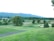 A lush, expansive golf course set against a backdrop of rolling hills and dense trees. The landscape features well-maintained grass, sand bunkers, and scattered trees, creating a serene and picturesque setting.