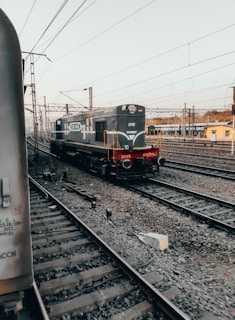 A railway yard with multiple tracks and an engine moving along one of them. Overhead electric lines are visible, along with a yellow building in the background. The scene is largely composed of steel and gravel, typical of a train station or railway hub.