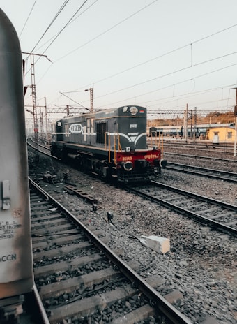 A railway yard with multiple tracks and an engine moving along one of them. Overhead electric lines are visible, along with a yellow building in the background. The scene is largely composed of steel and gravel, typical of a train station or railway hub.