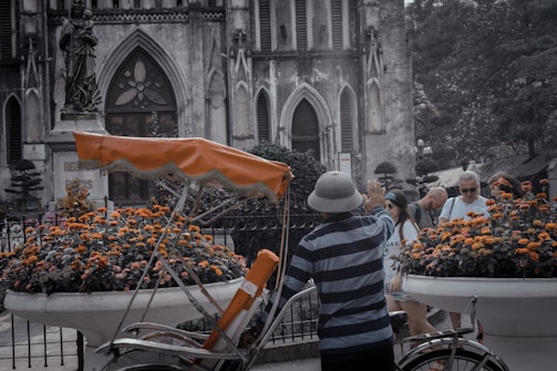 A vintage rickshaw is parked in front of a historic building with gothic architecture. The scene is accented by large flower pots filled with bright orange and yellow blooms. A person wearing a striped shirt and a hat is standing near the rickshaw, while a group of tourists, some wearing sunglasses, are visible in the background. The weathered stone facade of the building conveys an aged charm, with arched windows and intricate details.