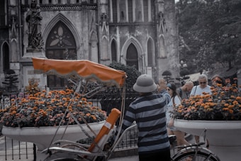 A vintage rickshaw is parked in front of a historic building with gothic architecture. The scene is accented by large flower pots filled with bright orange and yellow blooms. A person wearing a striped shirt and a hat is standing near the rickshaw, while a group of tourists, some wearing sunglasses, are visible in the background. The weathered stone facade of the building conveys an aged charm, with arched windows and intricate details.