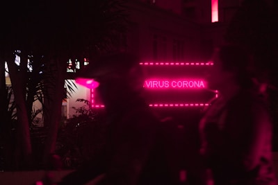 A neon pink sign displays the words 'Virus Corona' in a nighttime setting with silhouettes of people in the foreground. The environment is dark, with trees partially visible and a modern building in the background.